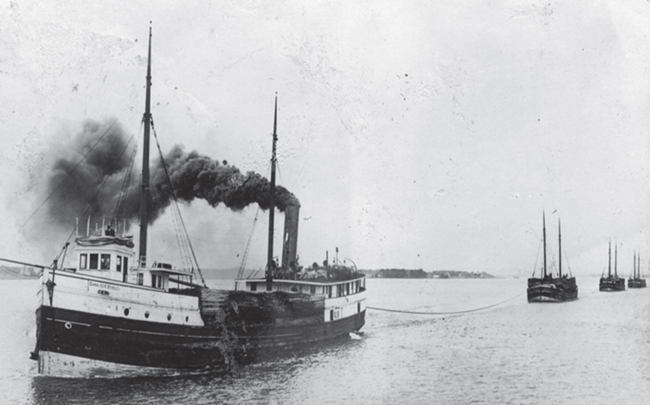 A boat, deck covered in stacks of cordwood, billowing black smoke, pulls a line attached to another boat. The line continues back, pulling another boat, and another.
