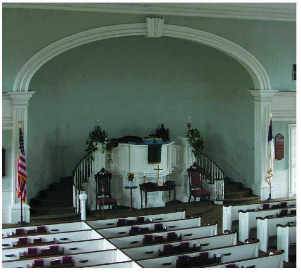 Two sets of stairs lead to the pulpit in set in a recessed half dome in a sanctuary with white walls, pews, and trim.
