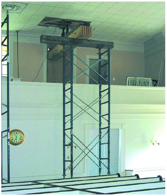 A beam atop scaffolding supports a crossbeam inside a white church room.