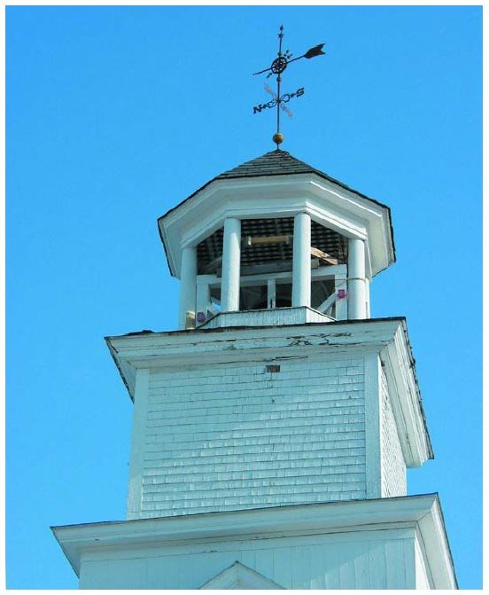 Weathered upper portion of a white four-sided steeple with lantern posts supporting a copula and a weathervane.