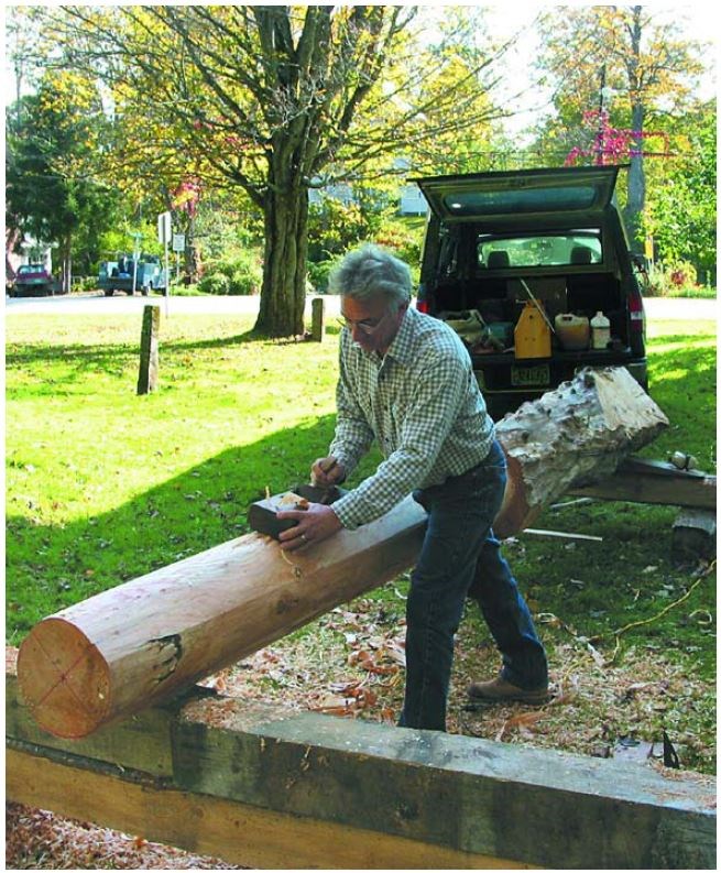 A white man with a hand planer smooths the edge of a large wooden post.