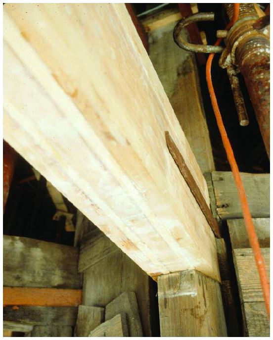 A thick horizontal pale colored beam connecting to a vertical post held in place with a iron bar.