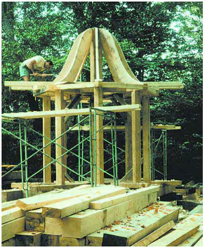 A man with an adze works on the timber framing of a cupola atop the lantern framework for a steeple. The framework is on the ground.