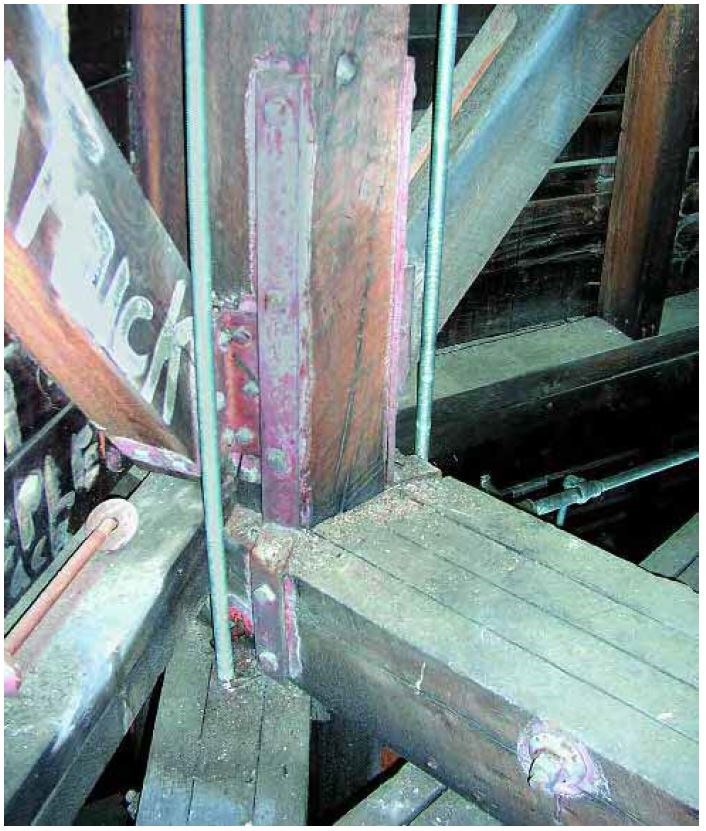 Iron straps and rods add support to a joint of five thick timber beams. Other framing in the background.