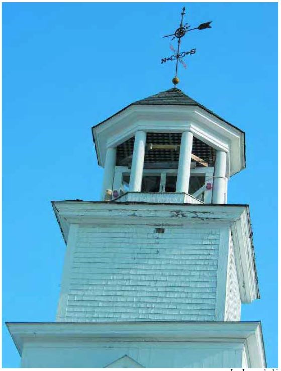 Weathered upper portion of a white four-sided steeple with lantern posts supporting a copula and a weathervane.