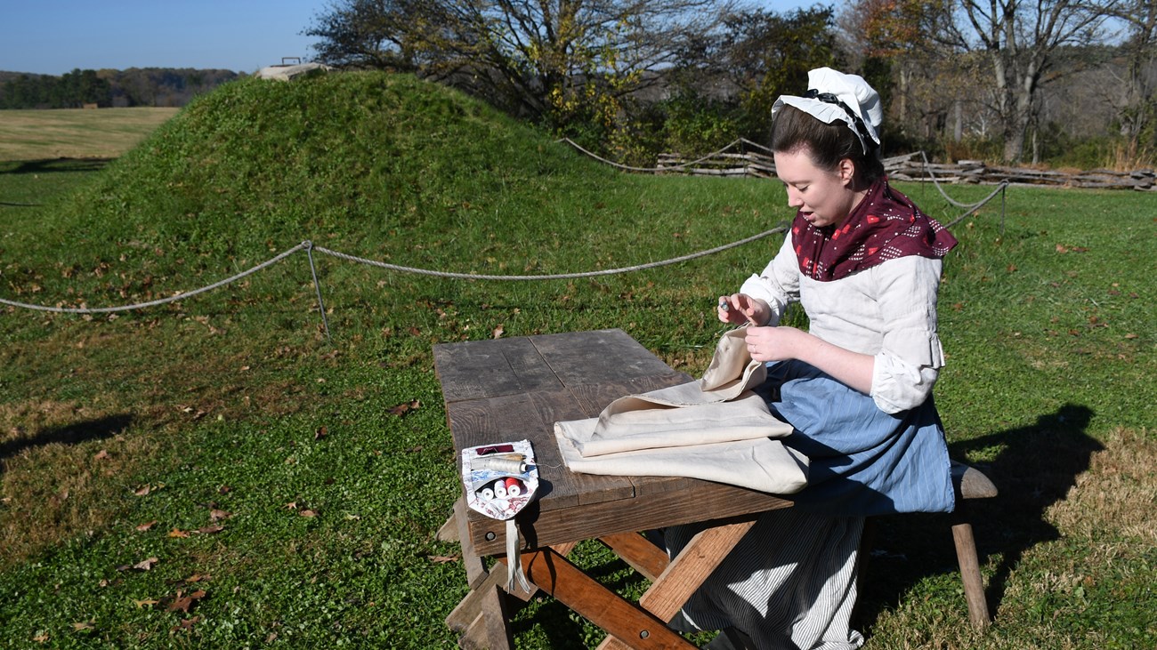 photograph, outdoors, wooden table, grass, woman sewing