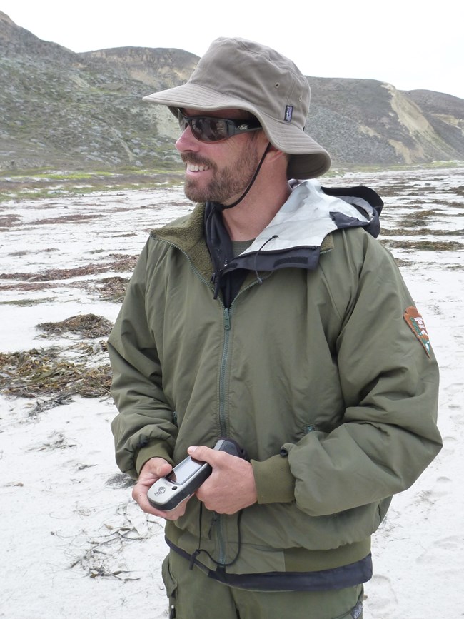 Steve holding a GPS on a sandy beach covered in patches of washed up kelp.