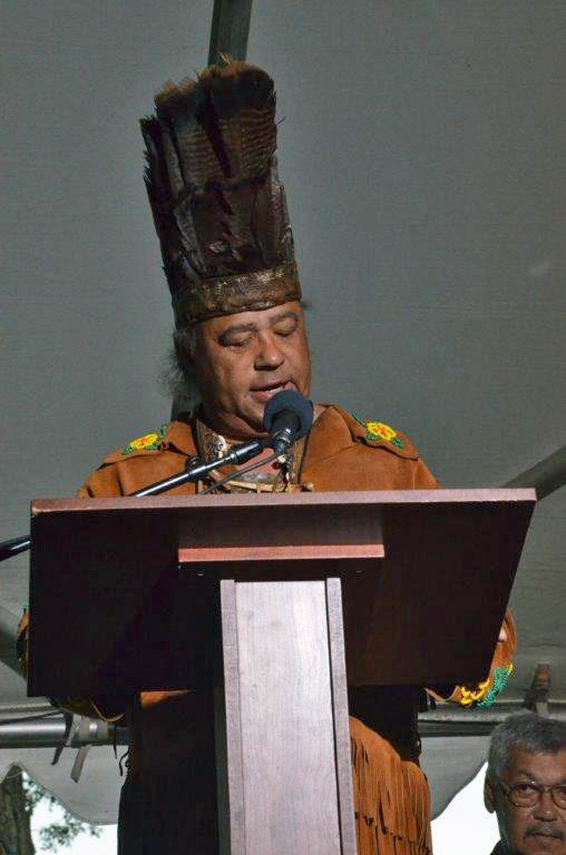 A man wearing a feather headdress and deerskin shirt speaks at a microphone under a tent