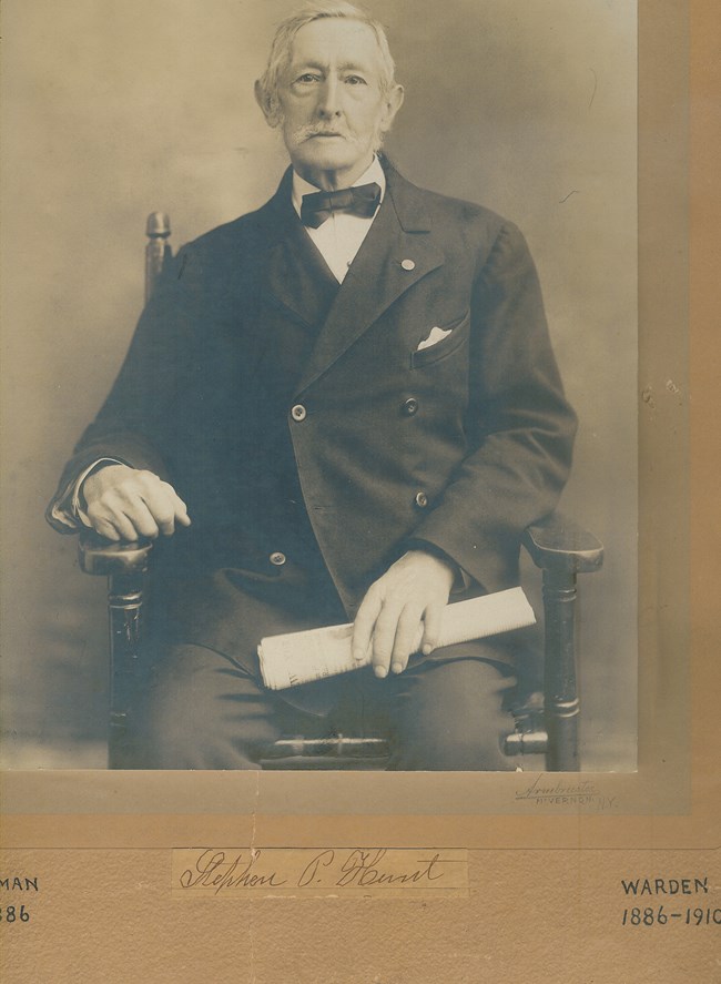 Man, seated, with mustache, dressed in suit, with bow tie, holding a paper