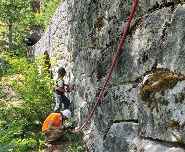 Two people in construction hard hats working on a stone support wall of a road running through a forest.