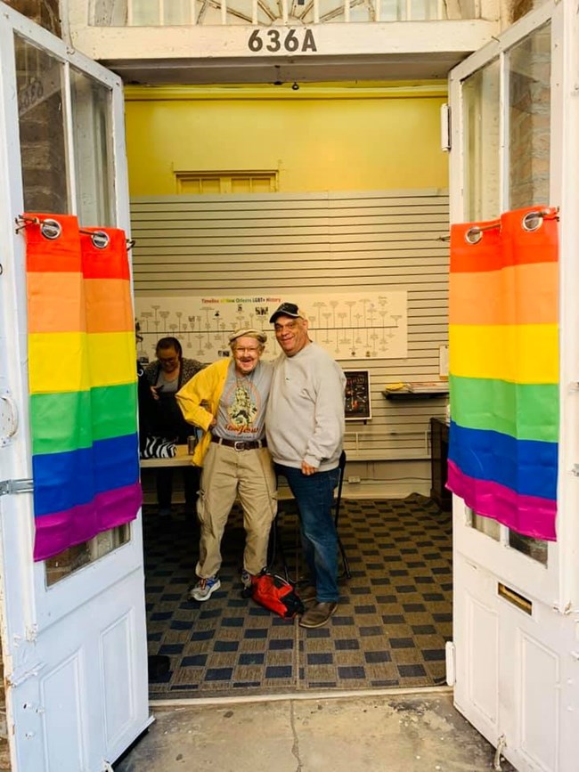 Two men standing and smiling at the camera with rainbow curtains on the windows on either side