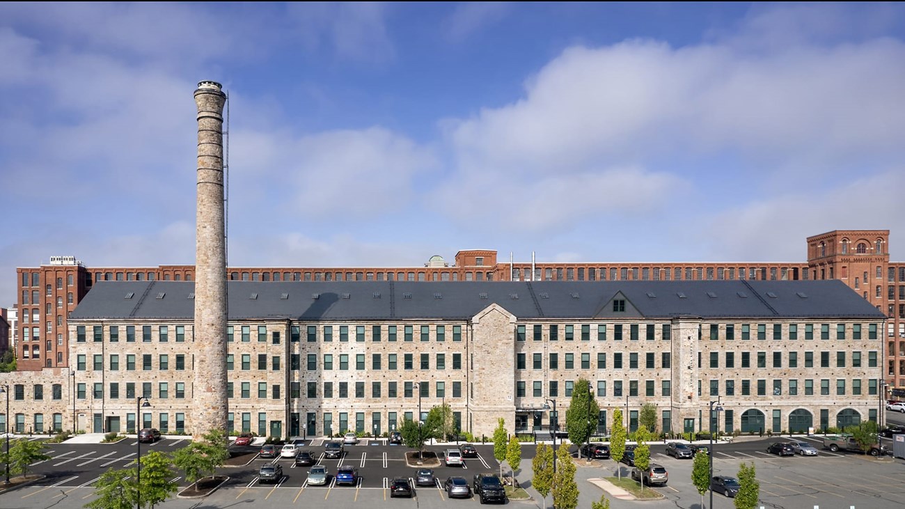 Large stone building made of brick with many windows along its side. Former stone mill, smokestack remains.