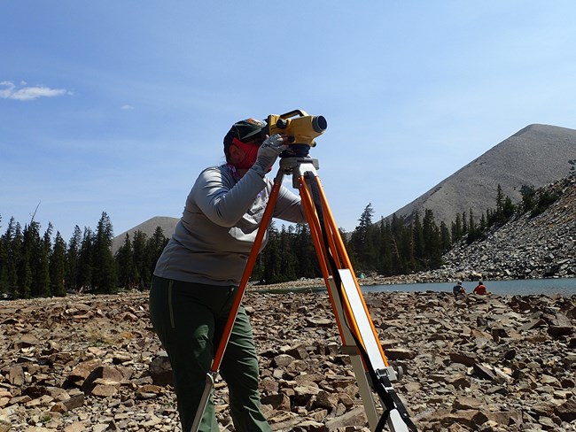 Woman in National Park Service field clothes stands on rocky lake shore and peers through a digital level logger scope to measure water level of a lake.