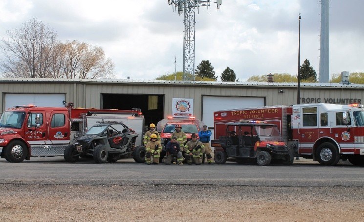 Two fire trucks and a group of people in yellow uniforms, outside a single-story flat roofed fire department building.