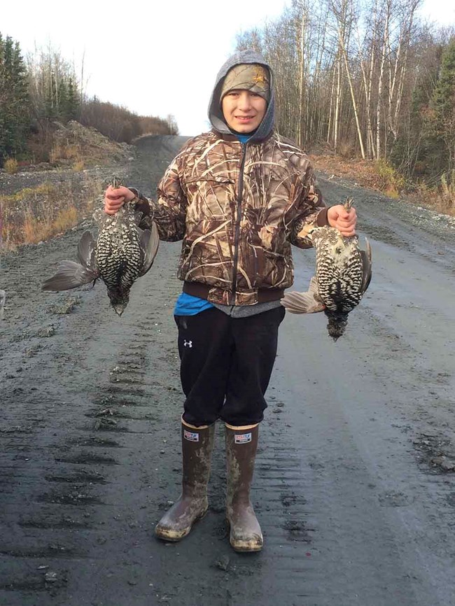 A boy holds a grouse in each hand.