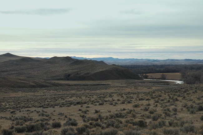 Sagebrush covered hills rise above the Little Bighorn River. Trees and mountains can be seen in the distance.