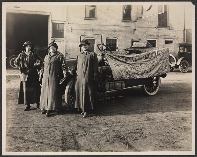 Suffrage Envoys Photograph of three women in coats and hats standing in front of automobile with banner