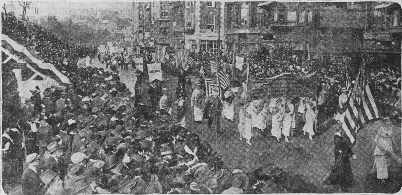 SuffrageParade-BG-Oct17-1915 crowds line the sidewalk as suffragists in white march down the street.