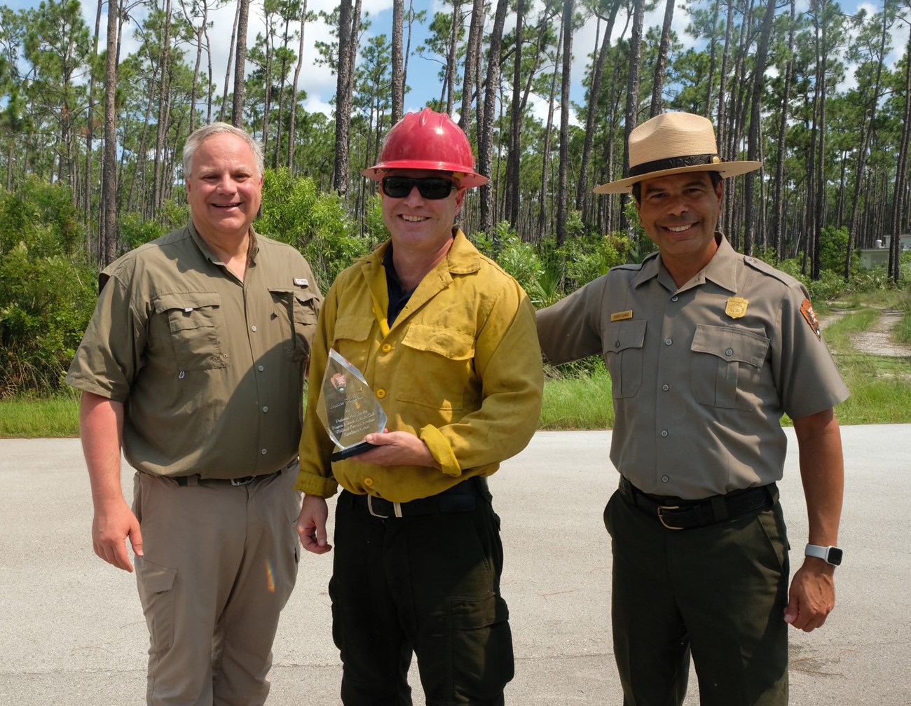 James Sullivan Receives Wildland Fire and Aviation Excellence Award Three men pose standing on asphalt in front of a forest. The man in center is holding an award.