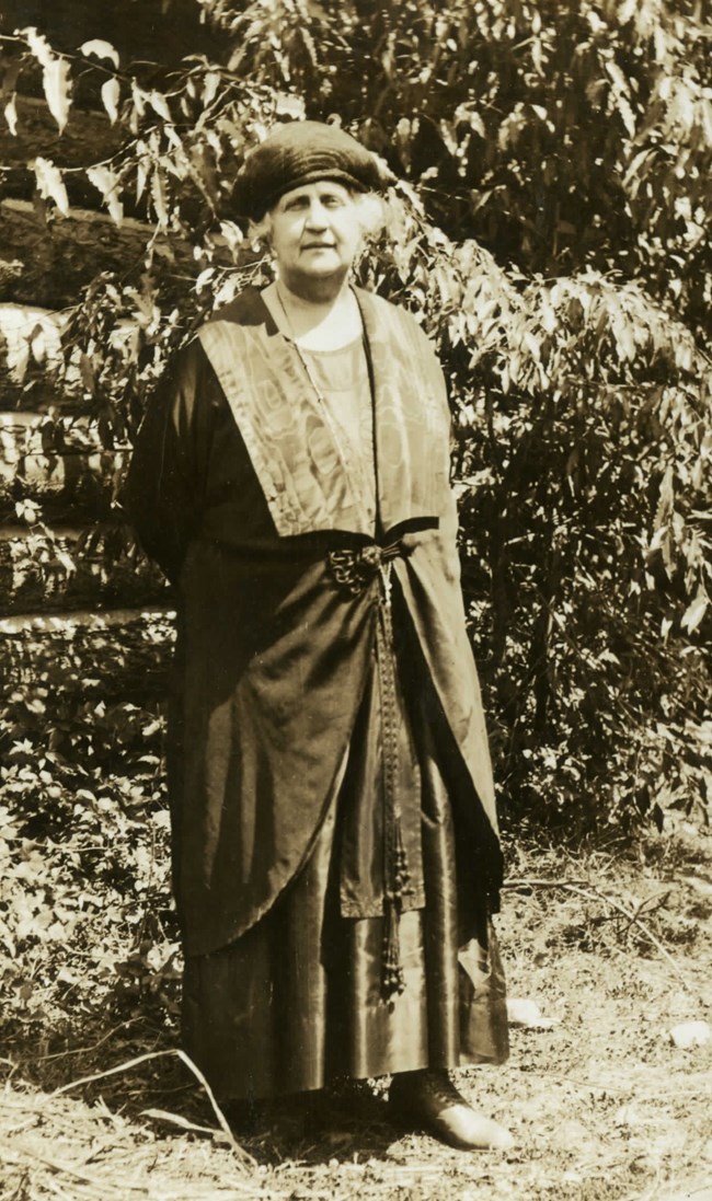 Mary Sullivan standing in front of a log building wearing a long dress.