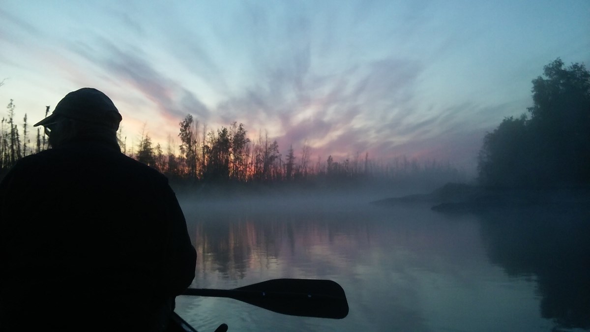 A man in a canoe holding a paddle is silhouetted against a river at sunrise with trees on the far shore.