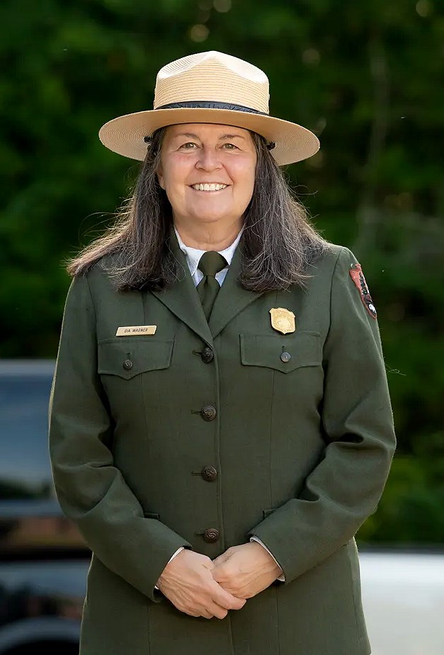 A smiling person with shoulder length brown hair, in a dark green, military-style uniform and a wide-brimmed tan colored hat.