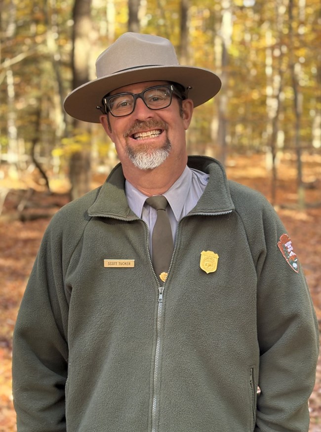 Smiling man in NPS uniform with NPS flannel jacket, straw hat, badges, eyeglasses, with hands in pockets in front of woodland in fall foliage