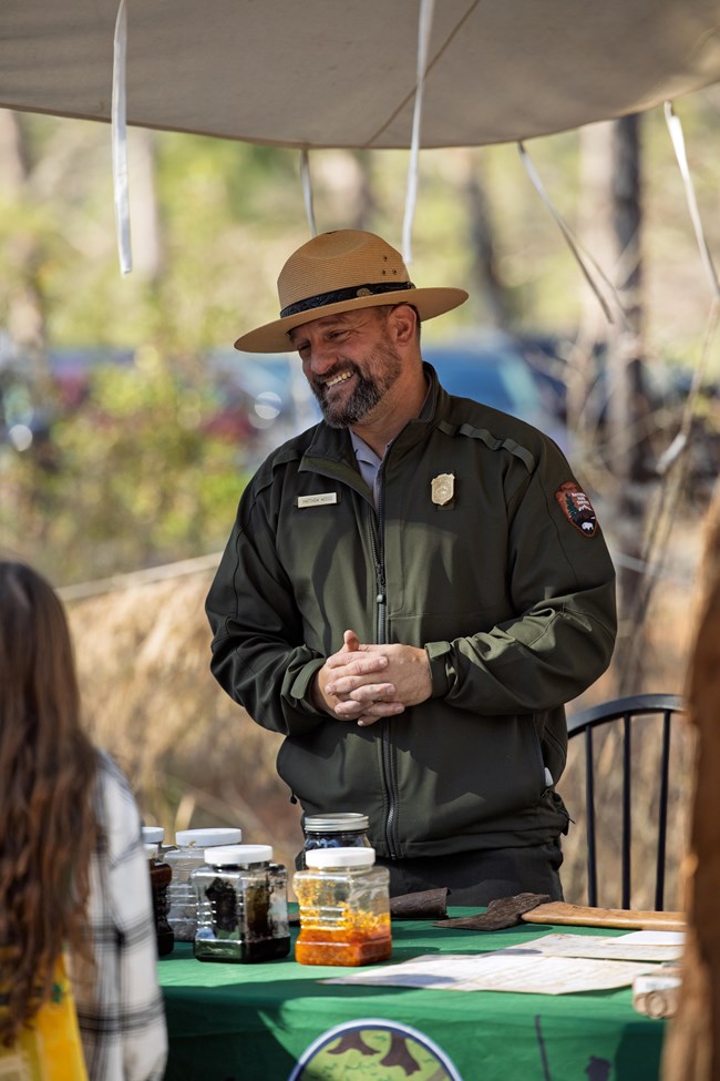A person in wide-brimmed straw hat, green jacket, smiles towards children across a table used to hold paint supplies.