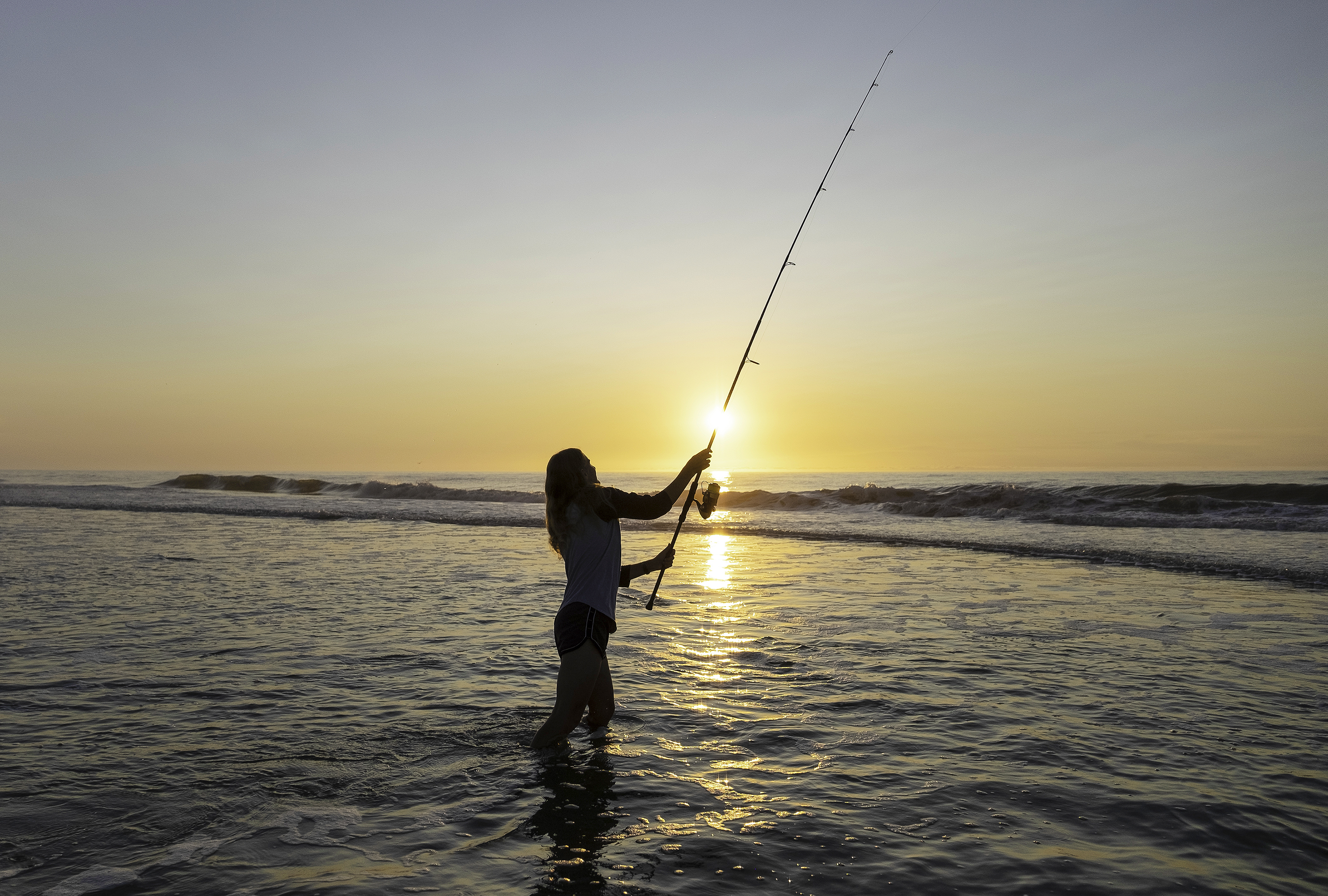 Woman with long fishing rod standing in ocean surf at sunset
