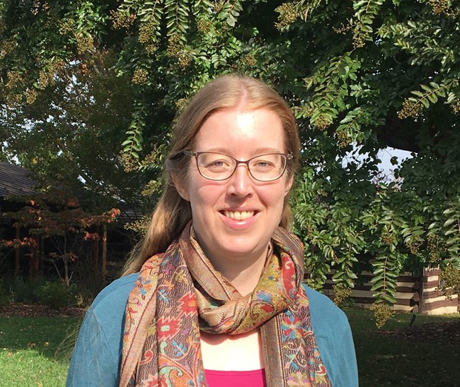 Suzy Traut a woman with straight brown hair and glasses stands in the sunshine under a tree.
