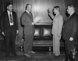 Newton Drury stands raising his right hand facing a man holding papers in his hand. Two other men look on.