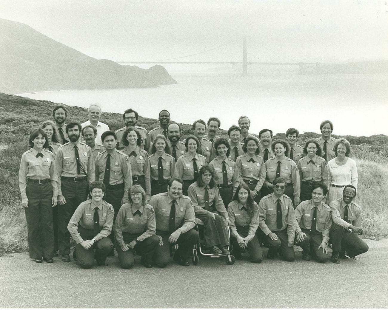 Group of Park Service employees in uniform pose in four rows  for a group photo with the Golden Gate Bridge in the distance.