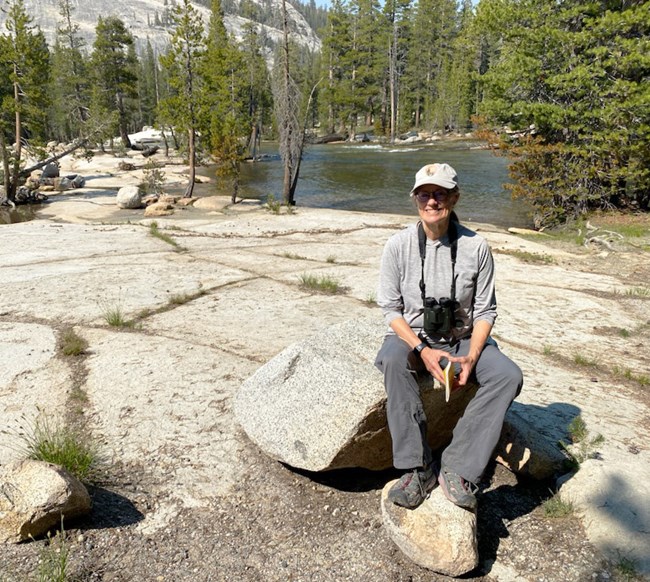 Sylvia Haultain, Tuolumne, Yosemite National Park Woman sits on granite boulder with river and conifer forest in background.