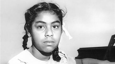 Black and white photo of a young girl with her dark hair in braids with one ribbon visible.