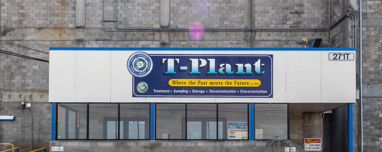 T Plant An image of a large sign on a concrete building reads “T Plant” with a blue background. Glass panes are directly below the sign.