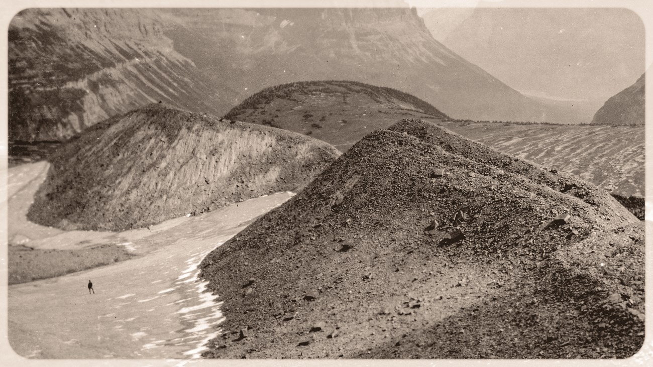 Black and white photograph of an alpine pass with a small glacier and a moraine in the foreground. A person is on the left in the distance. Black and white photograph of an alpine pass with a small glacier and a moraine in the foreground. A person is on the left in the distance.