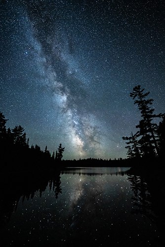 Milky Way reflected in water with tree silhouettes along each side