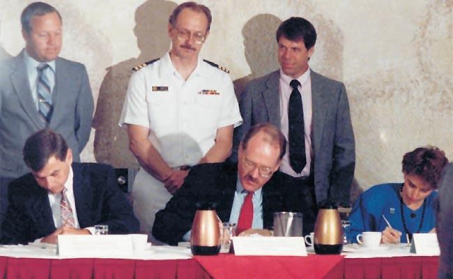Two men and a woman sign documents at a table while three men stand behind the seated group.
