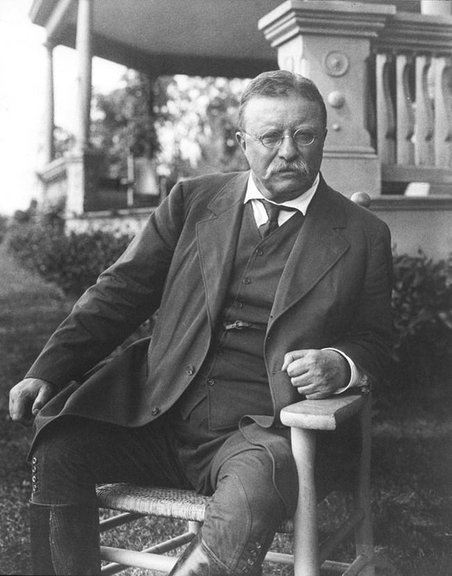A black and white photo of Theodore Roosevelt seated in front of the porch of his home, wearing a dark suit and riding boots.