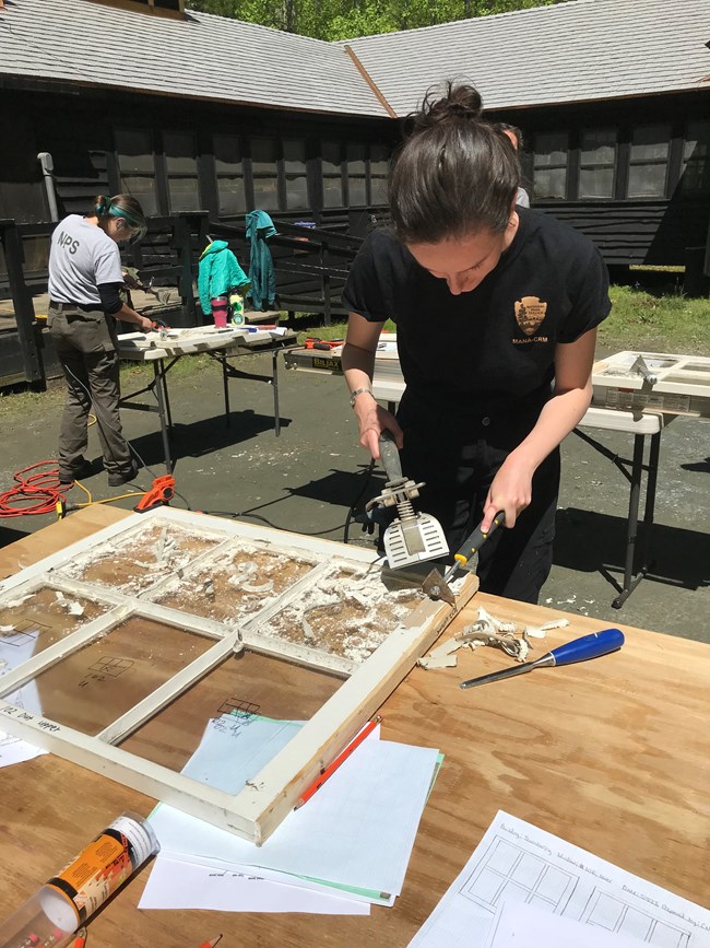 A person uses a heat gun and a metal scraper to remove old paint from a window frame on a worktable.