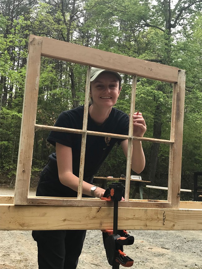 A person smiles through an open window frame that is clamped to a work bench.