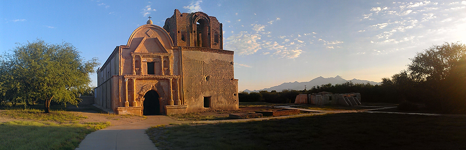 An old mission church next to adobe wall ruins next to paved paths and mown grass in a landscape of scattered trees and mountains in the distance.