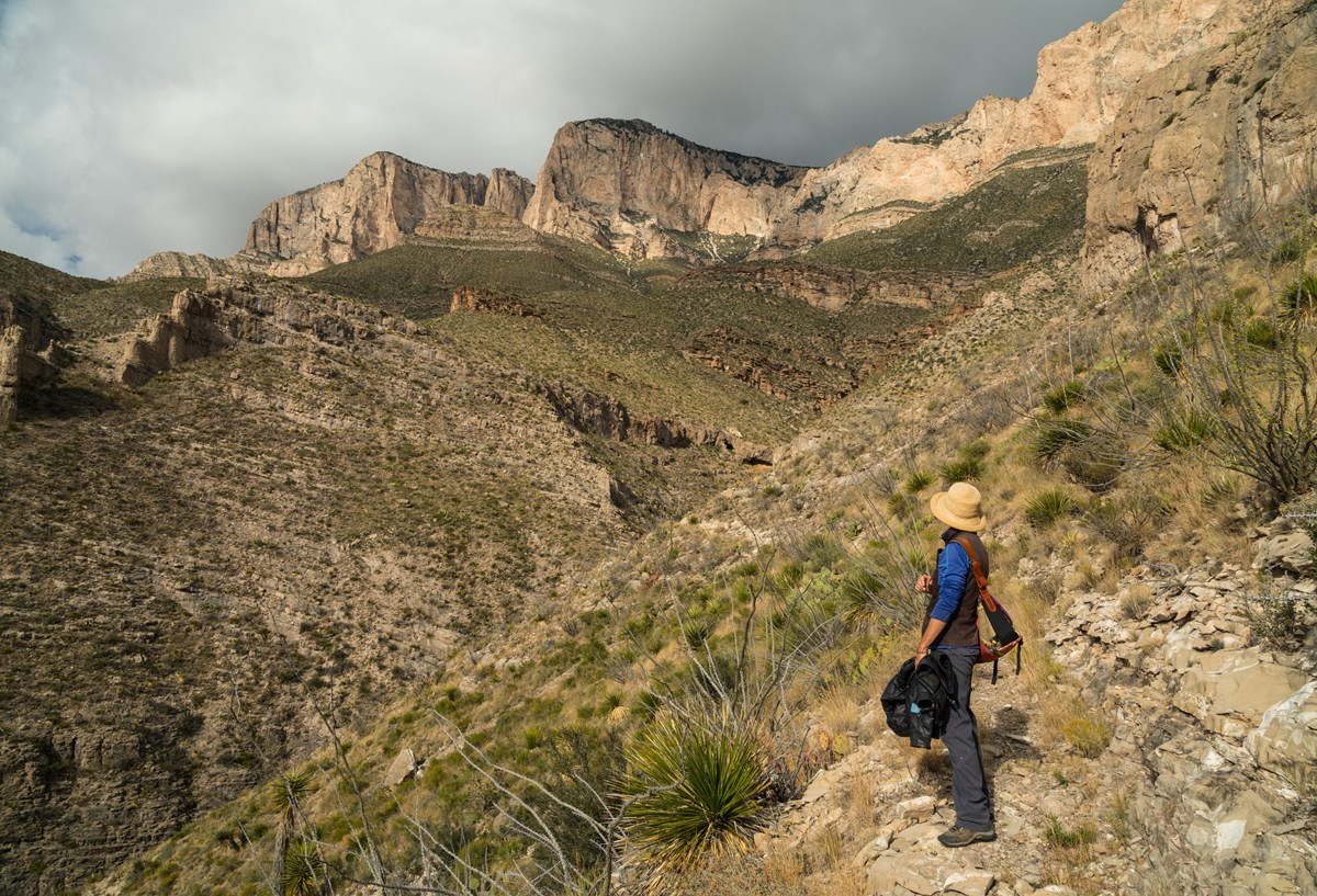 A hiker stands below tall and yellow desert mountain cliffs lit by the sun