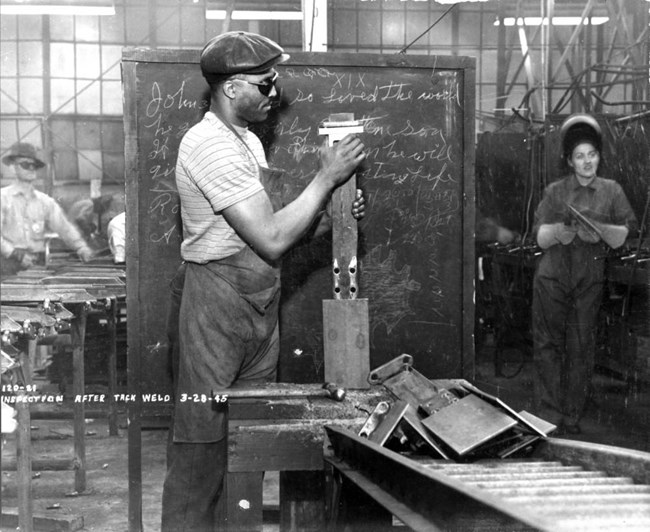 Black man wearing goggles and overalls inspects a long rectangular pillar of metal. Other pieces of welded metal lay on the conveyor belt next to him. He is standing in front of a chalkboard.