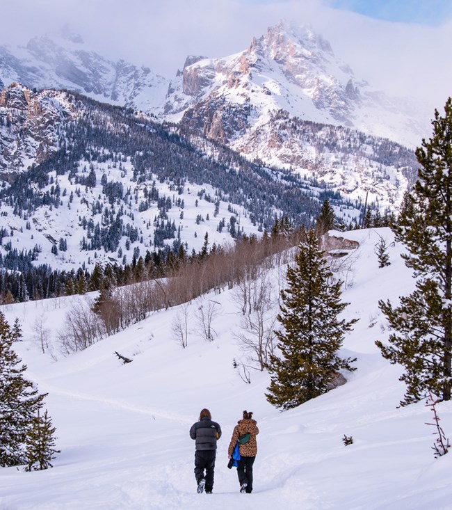Two visitors hike to Taggart Lake in the snow with the Teton range in the background and a snow-covered landscape.