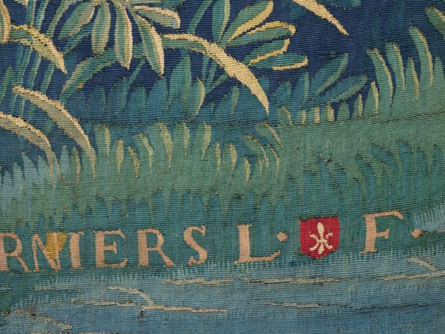 Detail of a tapestry with woven with the letters R N I E R S and a small shield flanked by the letters L and F.