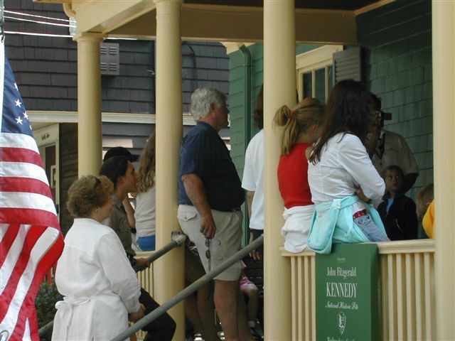 A group of Kennedy family members gather in front of a green colored house with cream colored banisters.  An American flag is on the far left of the photo.