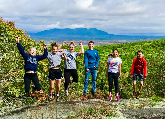 Teenagers do a jumping pose for a picture with a large forest and mountain in the background.
