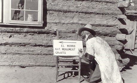 Elva Tharp kneels on the ground as she paints a sign in front of a log cabin. She is wearing a long robe over her uniform.