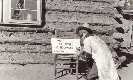 Elva-Sunshine-Tharp Elva Tharp kneels on the ground as she paints a sign in front of a log cabin. She is wearing a long robe over her uniform.